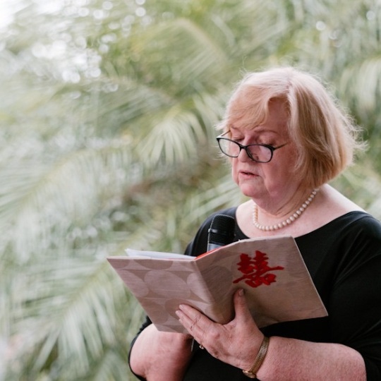 Jennifer Cram, Authorised Marriage
                              Celebrant reading from a gold brocade
                              folder that has a red Double Happiness
                              character on the front. She is holding a
                              microphone. She wears black clothing,
                              black framed eyeglasses, a string of
                              pearls and a gold bracelet