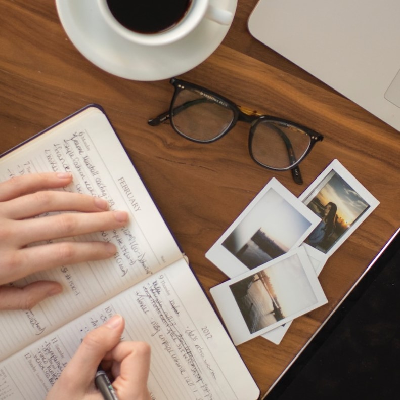 Hands
                      holding a pen and writing in a notebook. A cup of
                      coffee, a pair of eyeglasses and some photos are
                      nearby