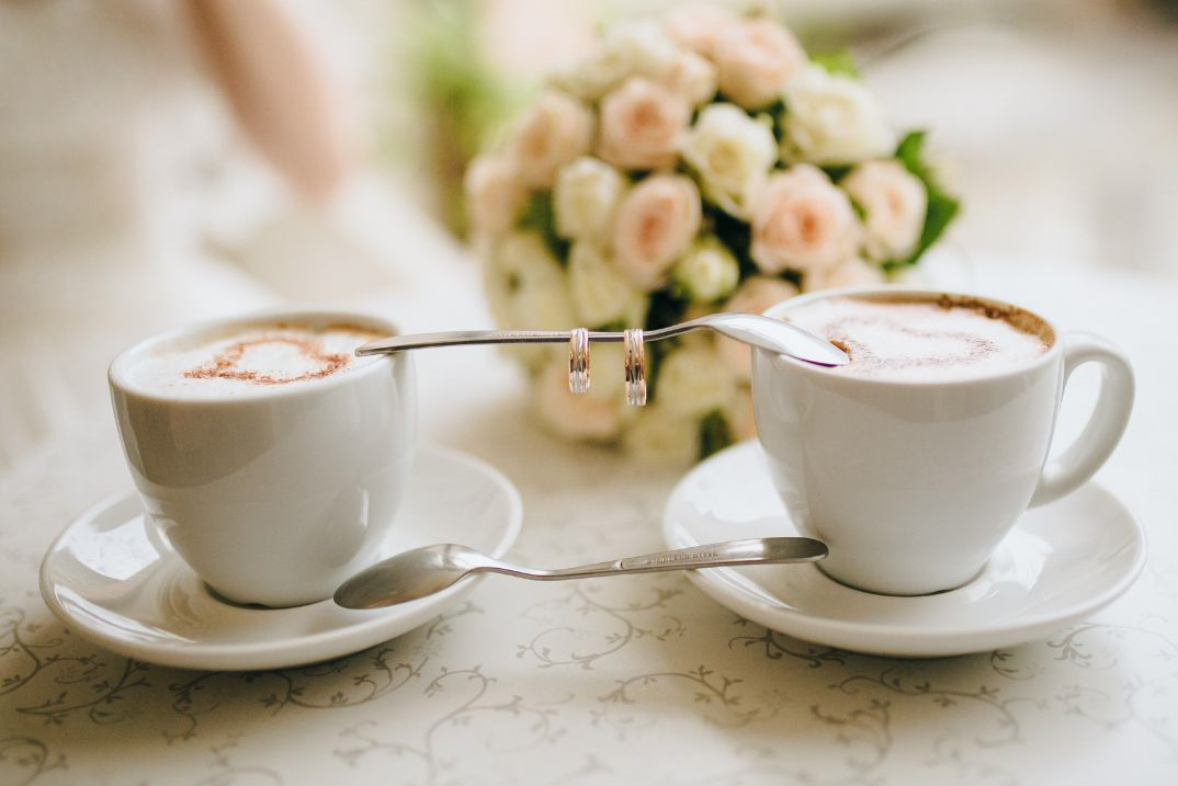 Two cups of
                  coffee with wedding rings hanging on the coffee spoons
                  and a wedding bouquet in the background