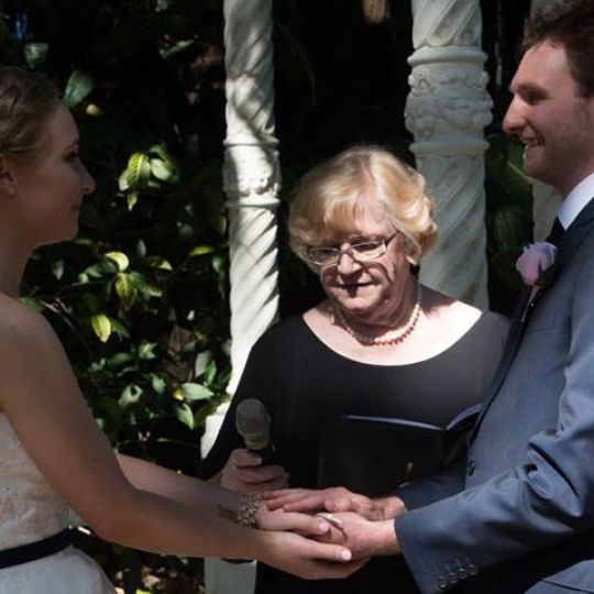 Jennifer Cram, Inclusive Marriage Celebrant
                      conducting a marriage ceremony. She is wearing an
                      understated black top and holds an unadorned A5
                      black folder and a black microphone. The bride and
                      groom are holding hands.