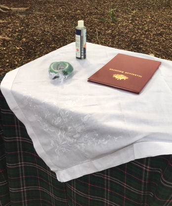 Signing Table with
                          Scottish National Tartan undercloth and white
                          on white embroidered linen topper cloth.
                          Maroon marriage register, green and white
                          handfasting cords, and a bottle of hand
                          sanitizer on the table.