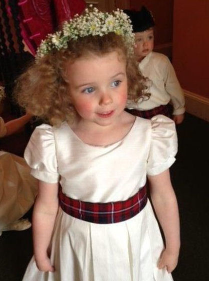 A flower girl wearing a white dress with a
                      tartan sash and a wreath of baby's breath on her
                      head
