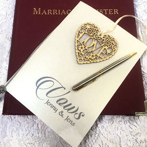 Cream card with the
                    words Vows Jenny & Jens on a burgundy marriage
                    register. Lying on the vows card is a wooden
                    filigree heart that includes the word love and a
                    silver pen lying on a pale carpet