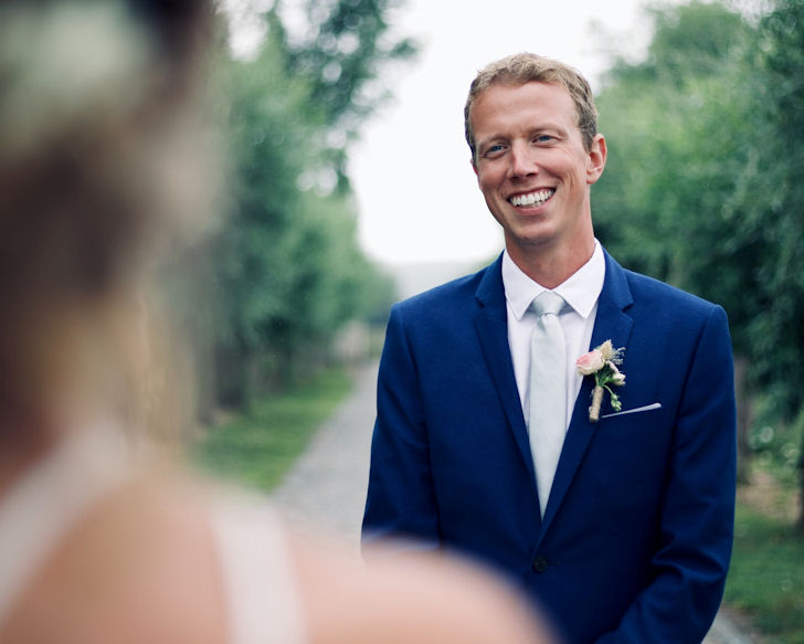 Bridegroom wearing
                        blue suit smiling at out-of-focus bride