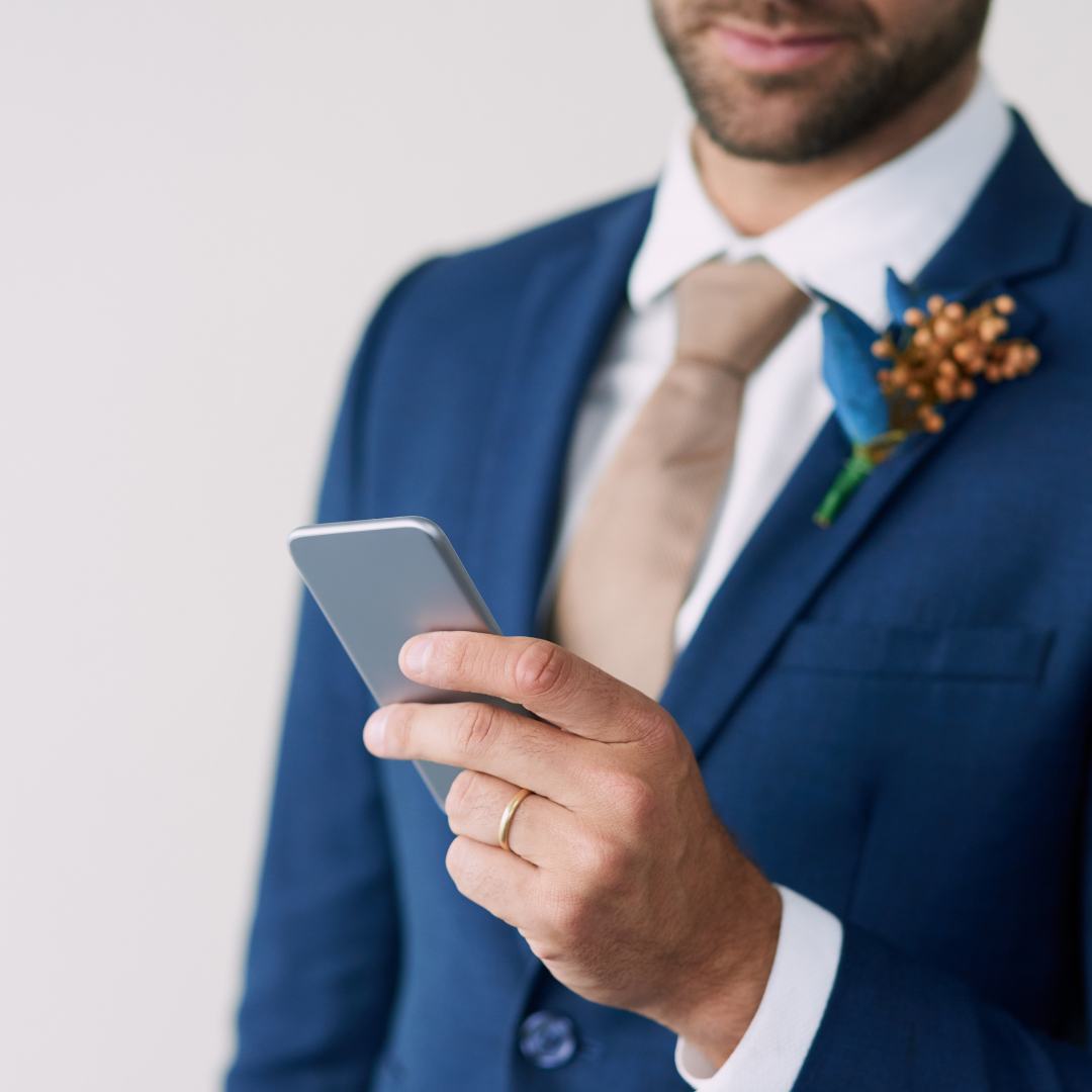 Groom
                      wearing a blue jacket on which is pinned a floral
                      boutonnier, is looking at the screen of a silver
                      smartphone