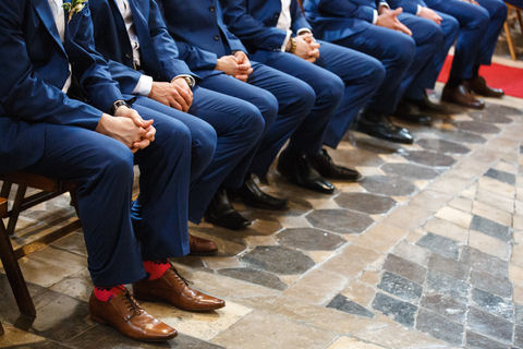 Groomsmen wearing blue
                        suits and brown shoes seated at front of wedding
                        ceremony