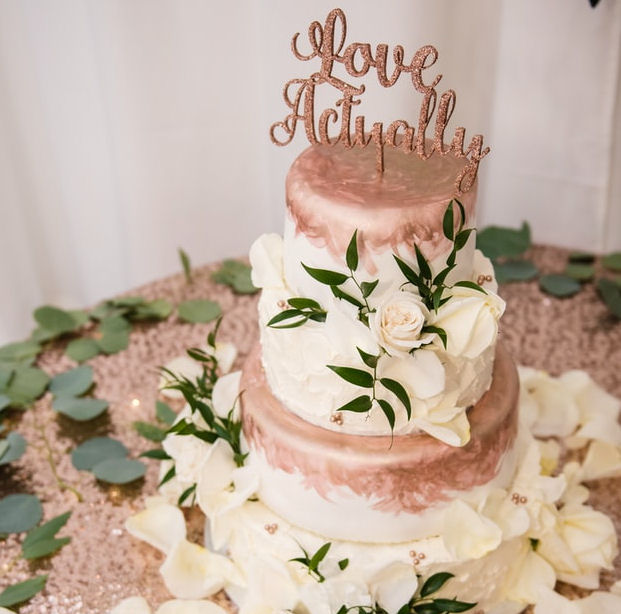 Wedding Cake with Love Actually topper
                      standing on a table covered in white rose petals
                      and green leaves