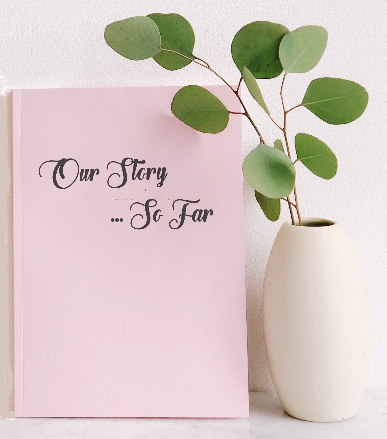 Pink Booklet with title Our Story ... So
                          Far upright next to white vase containing
                          green eucalyptus leaves