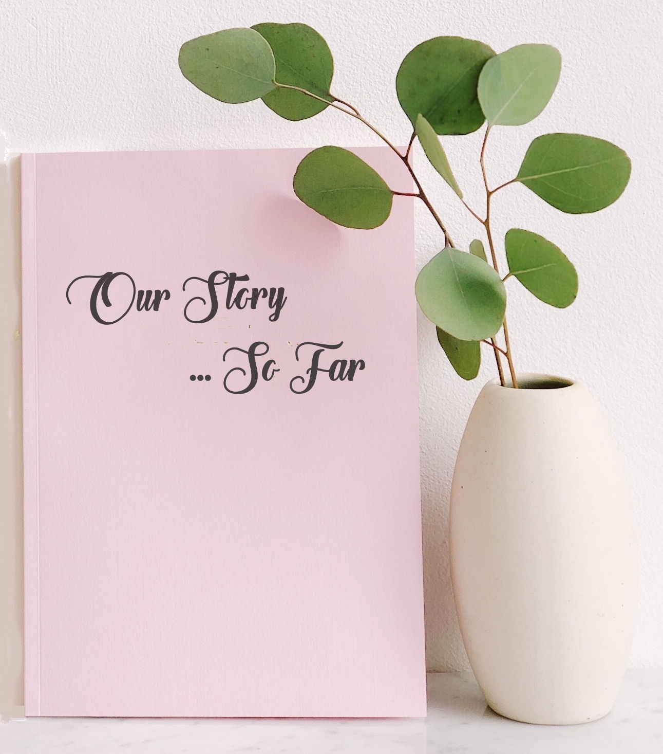 Pink Booklet with title Our Story ... So
                          Far upright next to white vase containing
                          green eucalyptus leaves