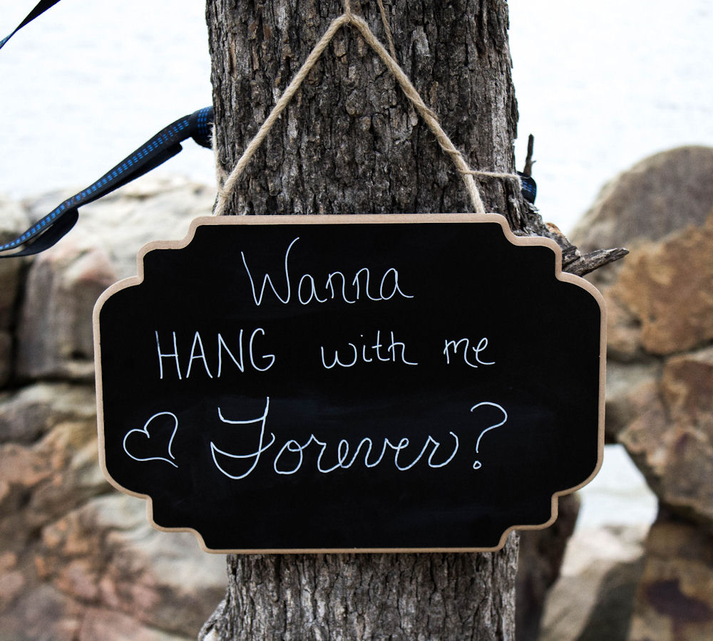 Chalkboard with "Wanna hang with me
                        forever" written on it, hanging on a tree.