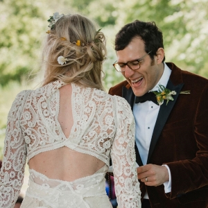 Groom laughing as he reads his vows. The
                    bride is seen only from the back. She is wearing a
                    lace dress with cut-outs.